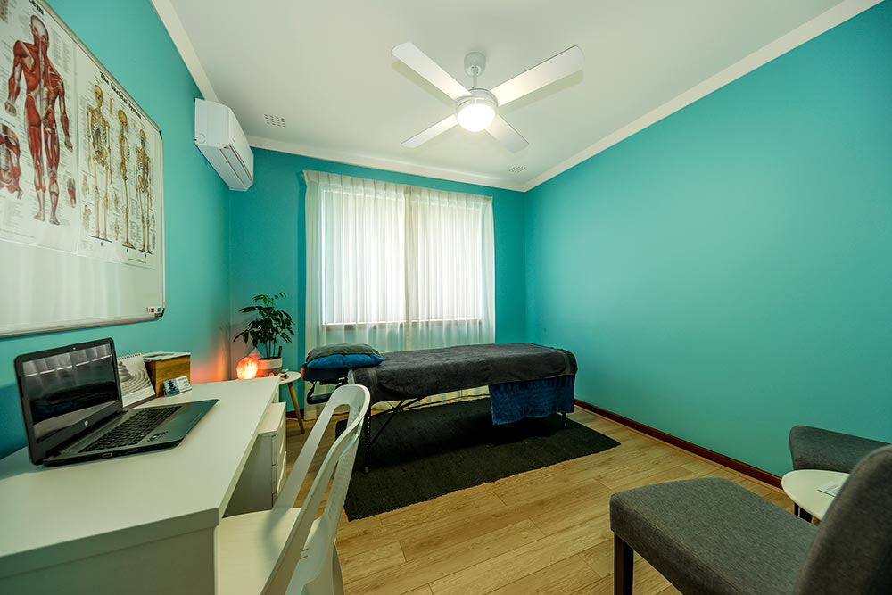 A teal blue Chiropractic room at Sarah's Circle with massage bed desk and chairs