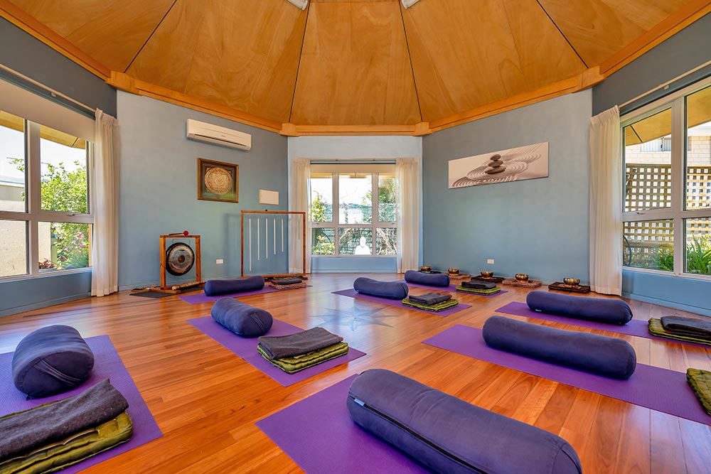 Large octagonal one room building with wooden floors and colorful purple exercise mats and cushions plus sound therapy instruments