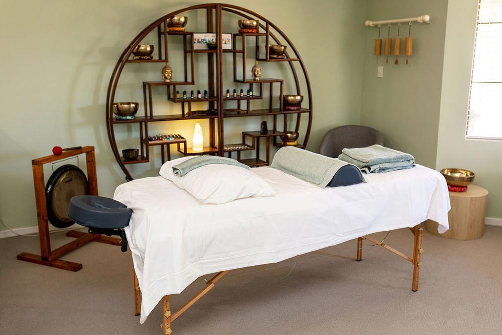 A Sound therapy room with massage table, gong, Tibetan Singing Bowl and large round wooden display cabinet filled with crystals and flower essences